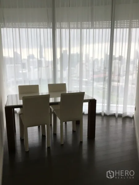 Condominium for rent in Pathum Wan, Bangkok - Dining nook with four white chairs and a glass-top table beside full-height sheer curtains and city views.