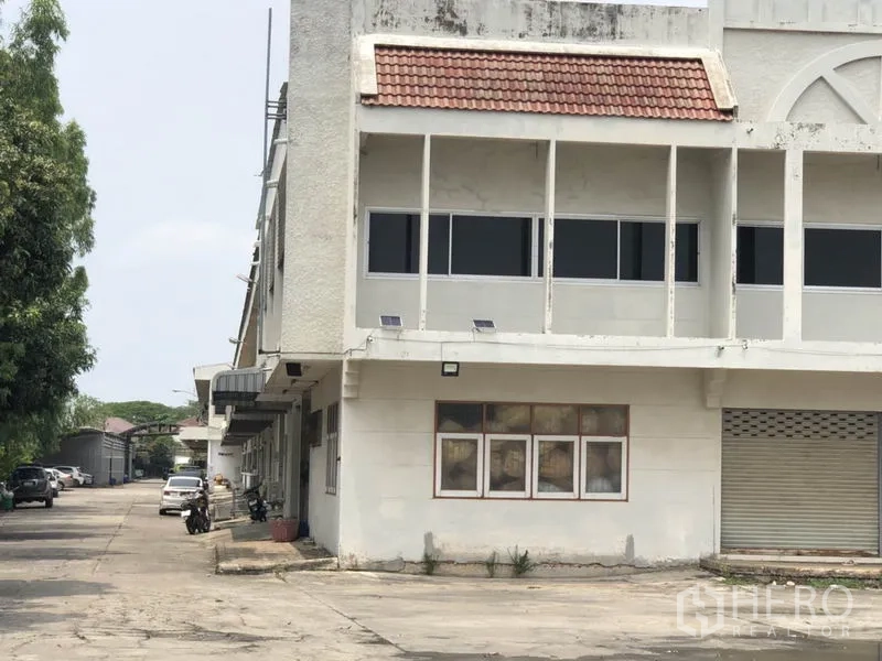 Warehouse for rent in Bang Kapi, Bangkok - Close view of the warehouse corner with shaded walkway, windows and a roller shutter beside a tree-lined driveway.