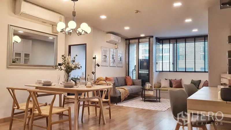 Condominium for rent in Khlong Toei, Bangkok - Bright open-plan living and dining area with wood floors, chandelier, large windows with blinds and a cozy sofa corner.
