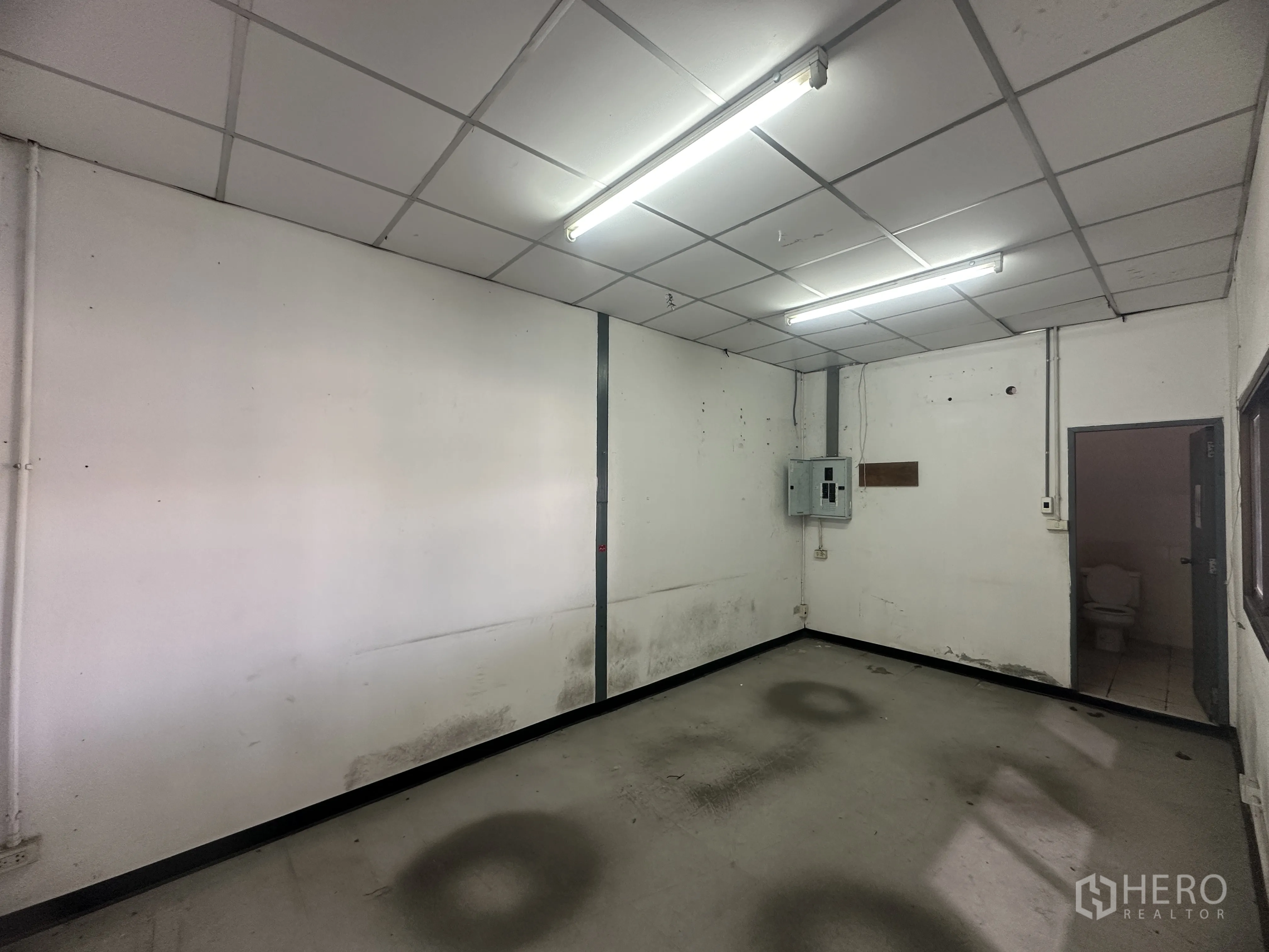Warehouse for rent in Khlong Luang, Pathum Thani - Simple office room with fluorescent lighting and a doorway to a restroom.