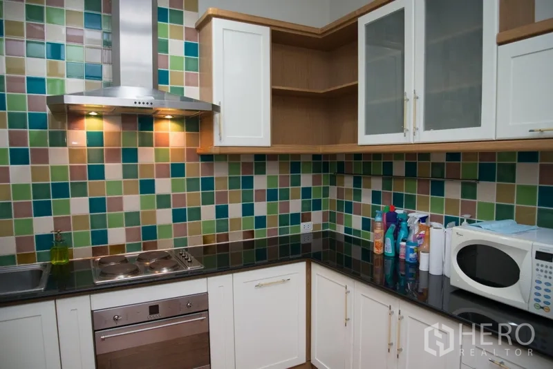 Condominium for rent in Khlong Toei, Bangkok - L-shaped kitchen featuring black stone countertops, oven, cooktop and white cabinetry.
