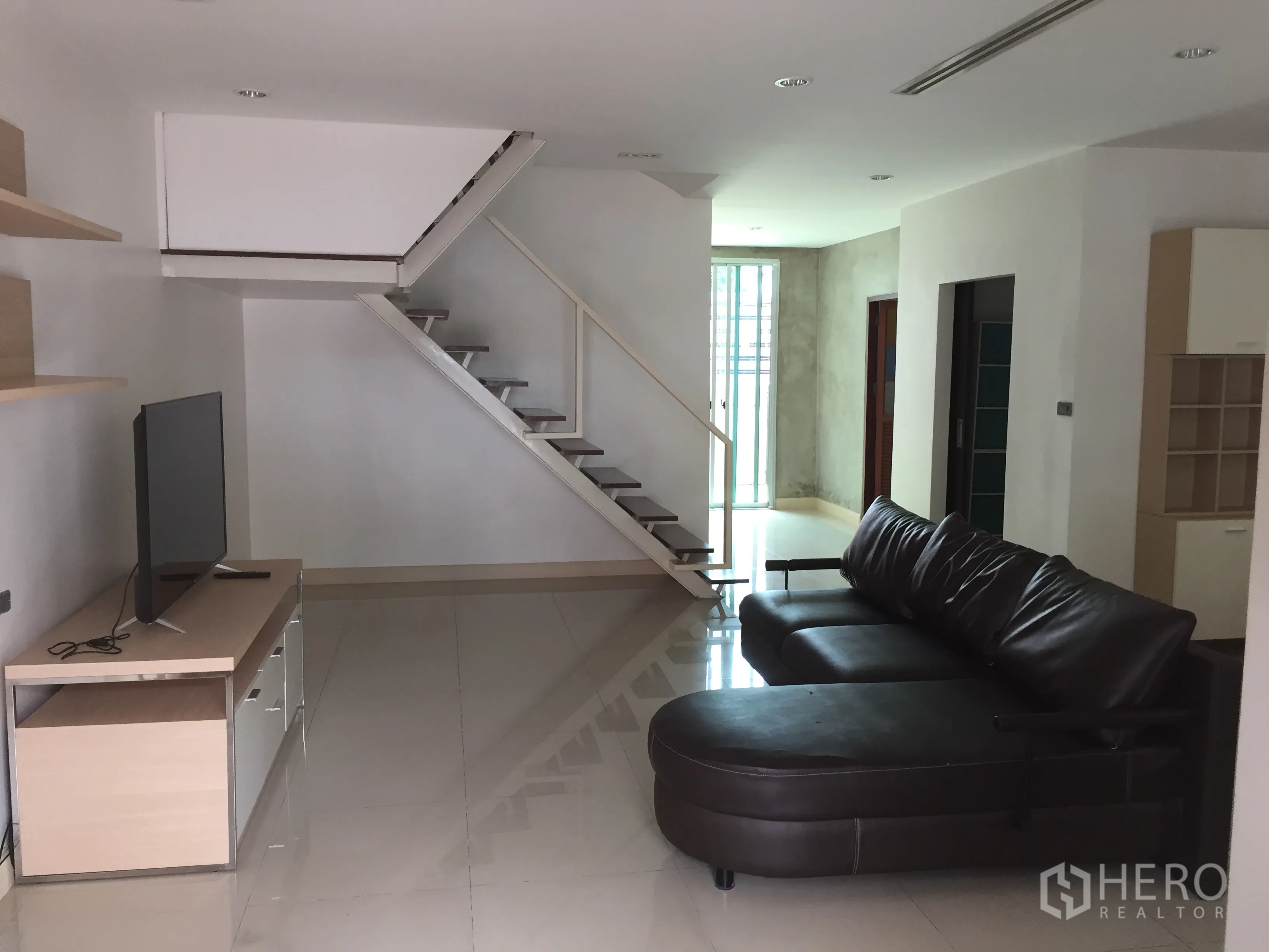 Townhouse for rent in Din Daeng, Bangkok - Open-plan living room with leather sectional sofa, TV console and a modern floating staircase.
