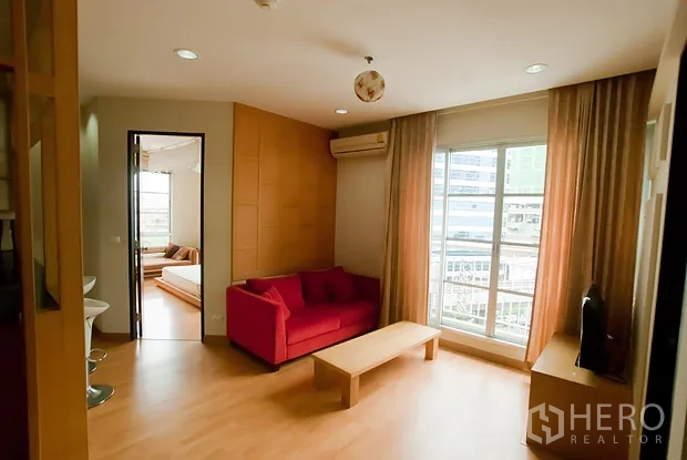 Condominium for rent in Khlong Toei, Bangkok - Bright living room with red sofa, wood flooring and large window facing a city-view balcony.