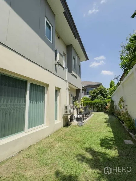 Detached House for rent or sale in Bang Khen, Bangkok - Side lawn and exterior wall of the two-storey home with windows and AC units.