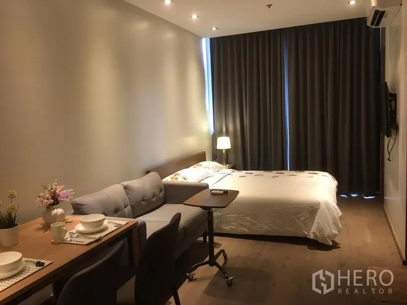 Condominium for rent in Khlong Toei, Bangkok - Open-plan living with dining table, sofa and bed facing tall drapes over warm wood flooring.
