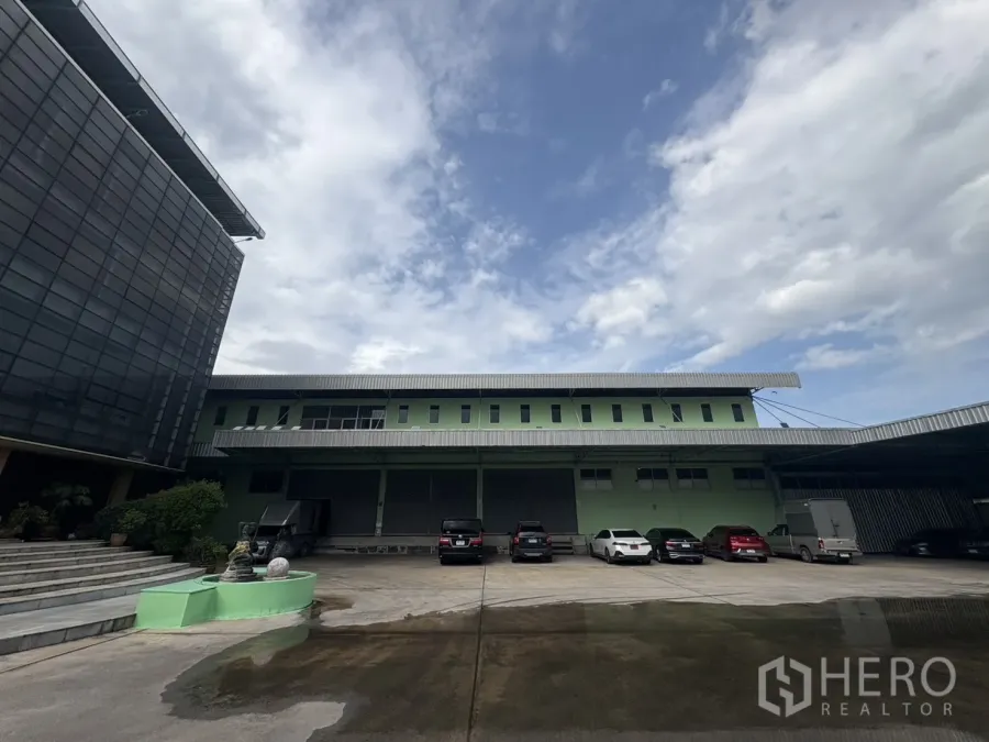 Warehouse for rent in Bang Khun Thian, Bangkok - Two-storey green warehouse exterior with covered loading bays, parking and an office block under a cloudy sky in Bang Khun Thian.