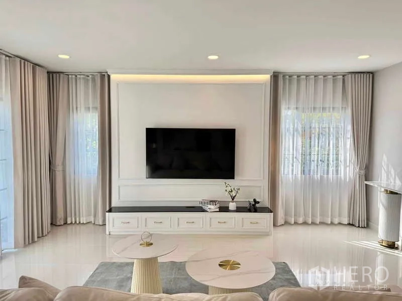 Detached House for rent or sale in Bang Phli, Samut Prakan - Living area with wall-mounted TV, long built-in console and large windows with sheer curtains.