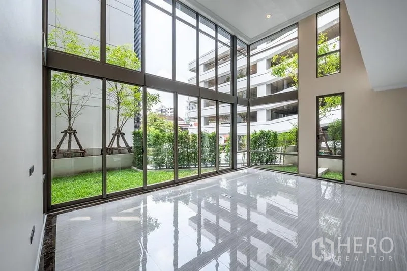 Townhouse for rent in Khlong Toei, Bangkok - Double-height living room with full-height glass walls opening to a private green garden.