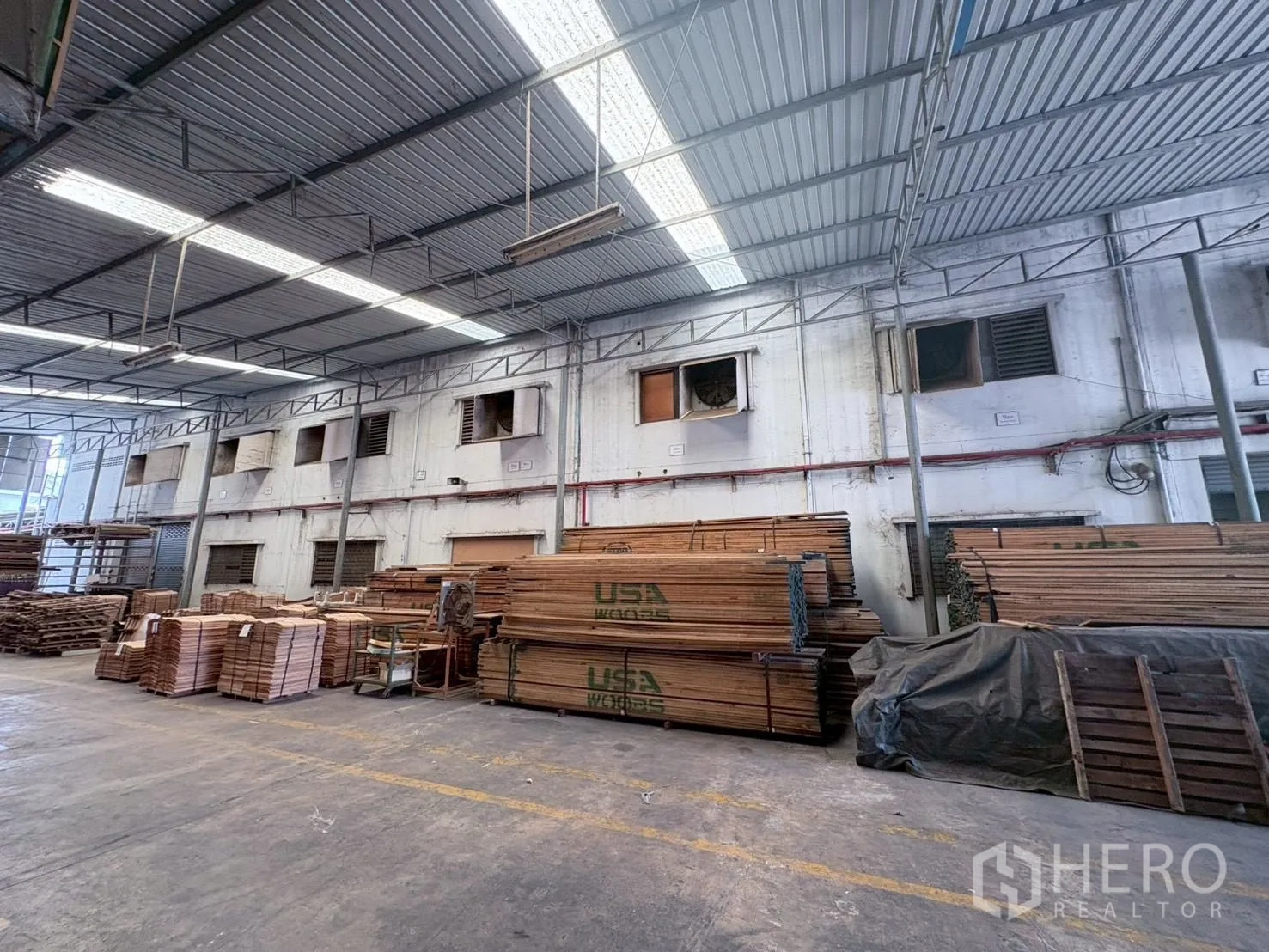 Factory for rent or sale in Bang Khun Thian, Bangkok - Covered warehouse area showing skylit metal roof and large stacks of timber.