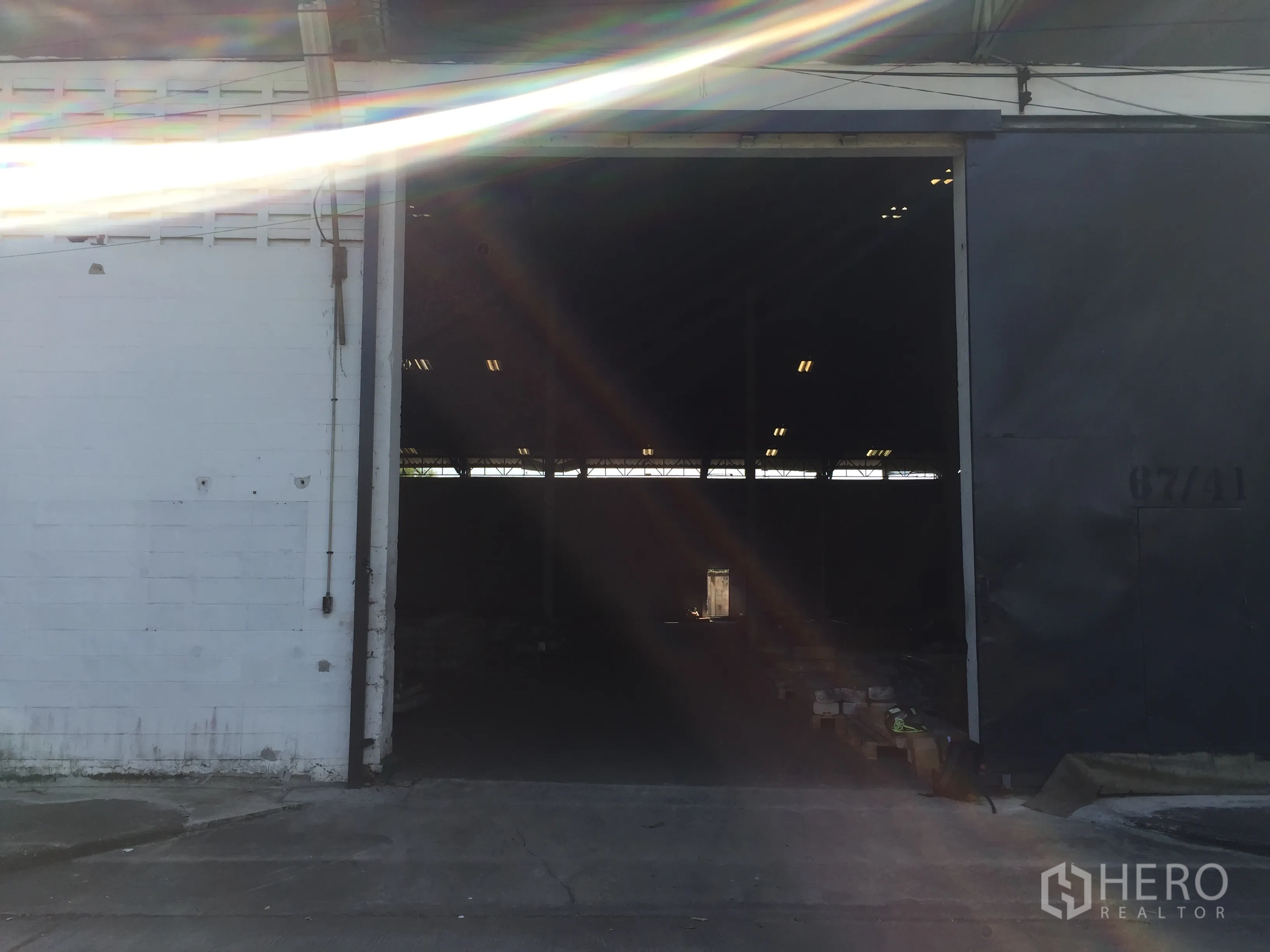 Factory for rent in Pathum Thani - Large sliding metal door opening to a dark warehouse bay with sunlight flare at the entrance.