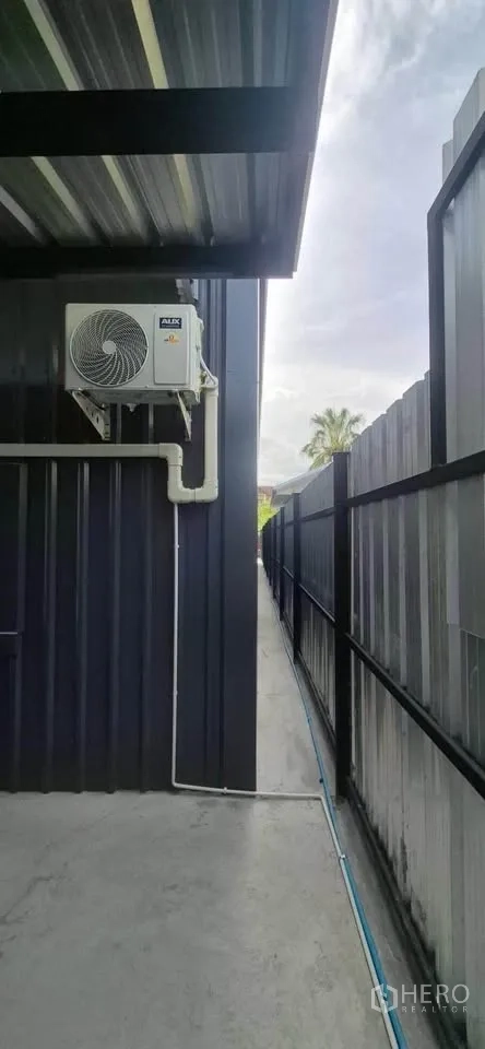 Warehouse for rent in Lat Phrao, Bangkok - Side walkway beside the warehouse with AC compressor, roof overhang, and metal fence.