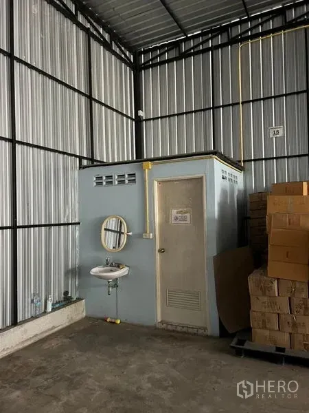 Warehouse for rent in Lat Phrao, Bangkok - Warehouse corner showing a built-in restroom with door and wall-mounted sink, exposed utilities, and cartons stored nearby.