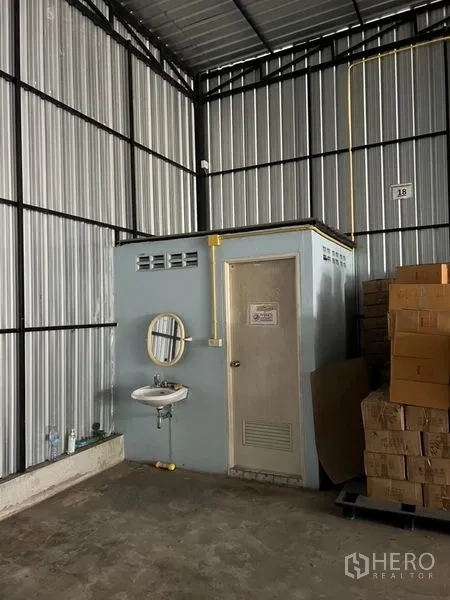 Warehouse for rent in Lat Phrao, Bangkok - Warehouse corner showing a built-in restroom with door and wall-mounted sink, exposed utilities, and cartons stored nearby.