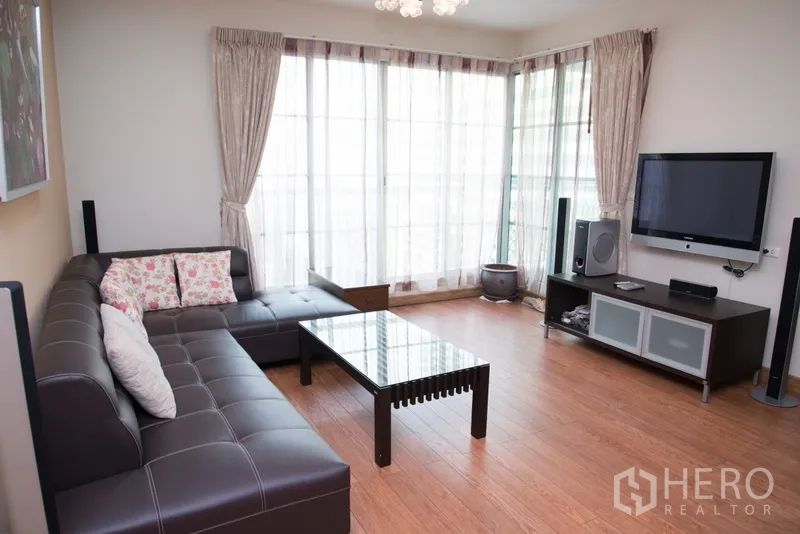 Condominium for rent in Khlong Toei, Bangkok - Living room with TV wall unit, surround speakers and large curtained windows providing natural light.