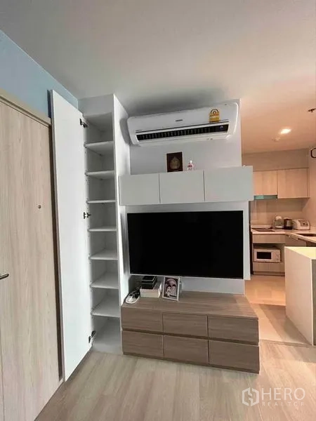 Condominium for sale in Pathum Wan, Bangkok - Modern living room with built‑in TV wall, open shelving, air conditioner, and light wood floors next to a compact kitchen.