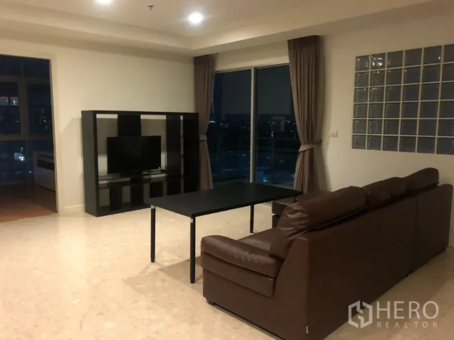 Condominium for sale in Khlong Toei, Bangkok - Spacious living room with brown leather sofa, TV console and large windows showing the night city view.