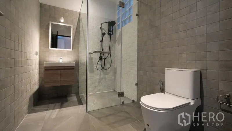 Townhouse for rent in Khlong Toei, Bangkok - Contemporary bathroom featuring a glass-enclosed shower, wall-mounted vanity, and gray tiles.