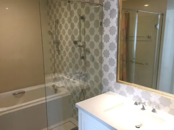 Condominium for rent or sale in Khlong Toei, Bangkok - Modern bathroom with glass shower over bathtub, patterned tile wall and white vanity with mirror.