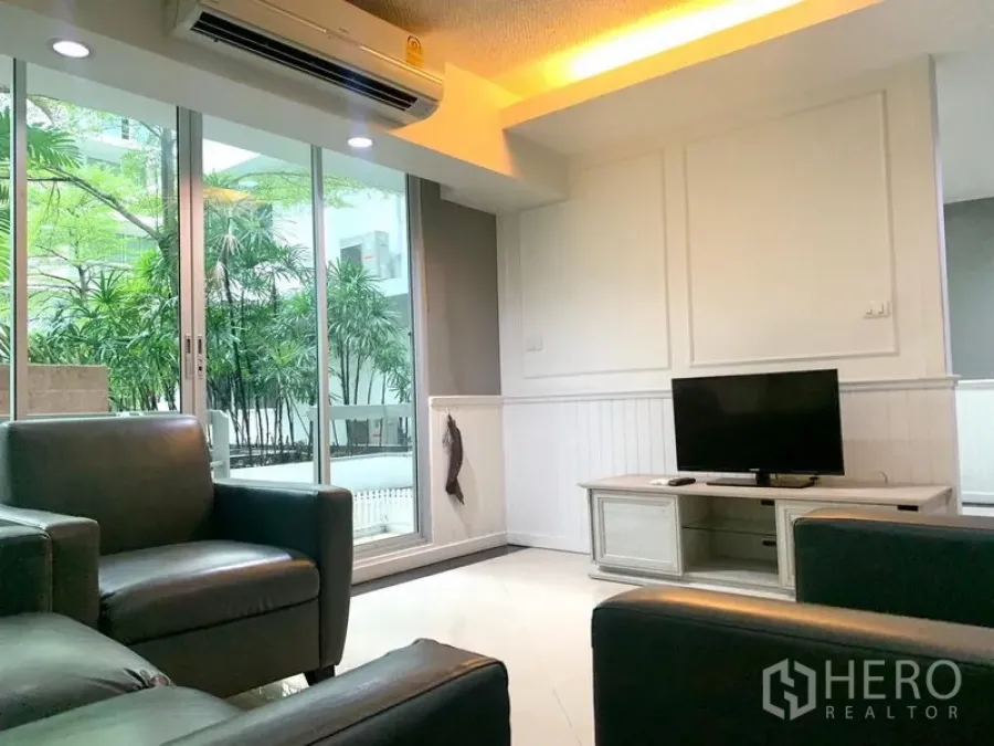 Condominium for rent in Khlong Toei, Bangkok - Living room with TV console, leather seating and sliding glass doors to garden-facing balcony.