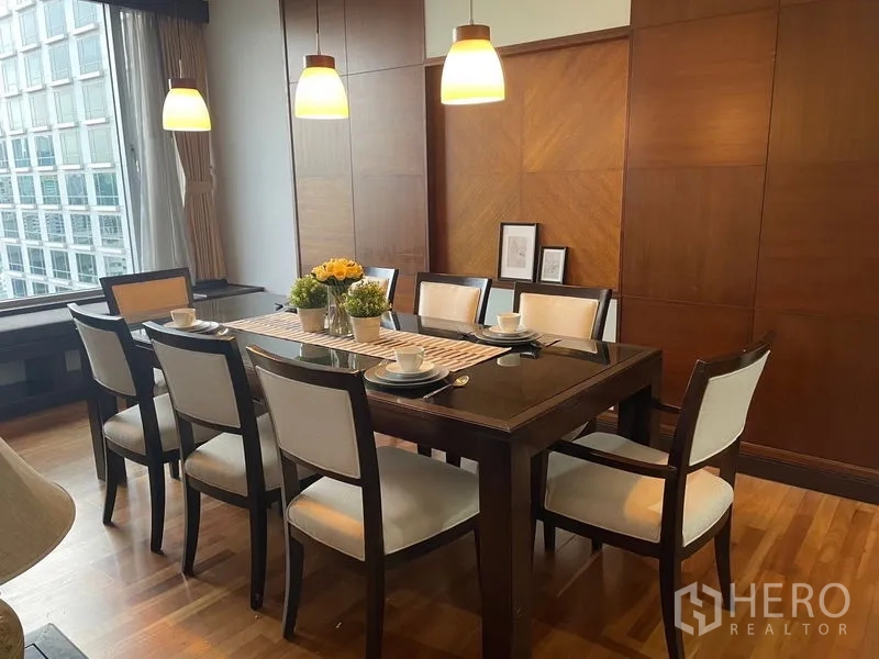 Condominium for rent in Pathum Wan, Bangkok - Formal dining room with glass-top table, wooden paneled wall and warm lighting.