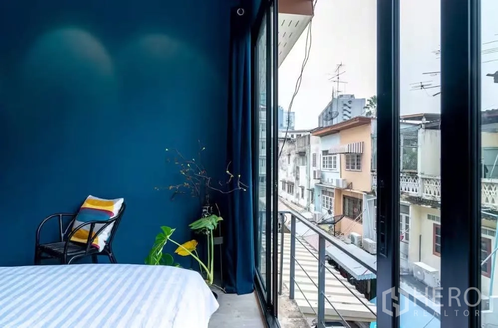 联排别墅 for rent in Watthana, Bangkok - 卧室一角，深蓝色墙面，椅子，落地推拉玻璃门通向小阳台眺望街景。