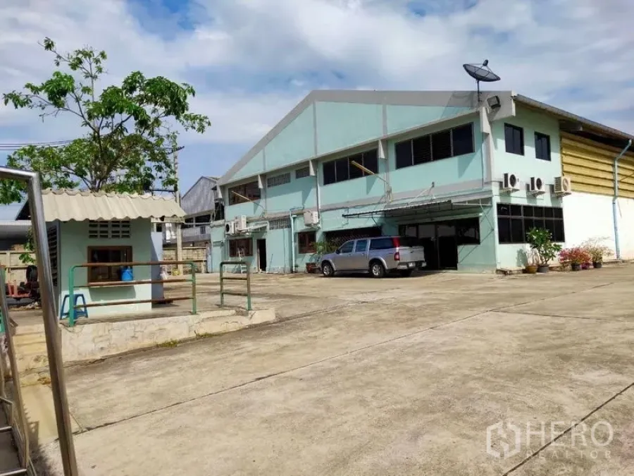 Warehouse for rent or sale in Bang Bon, Bangkok - Front view of a large two‑story aqua warehouse with a wide concrete yard and a pickup truck.