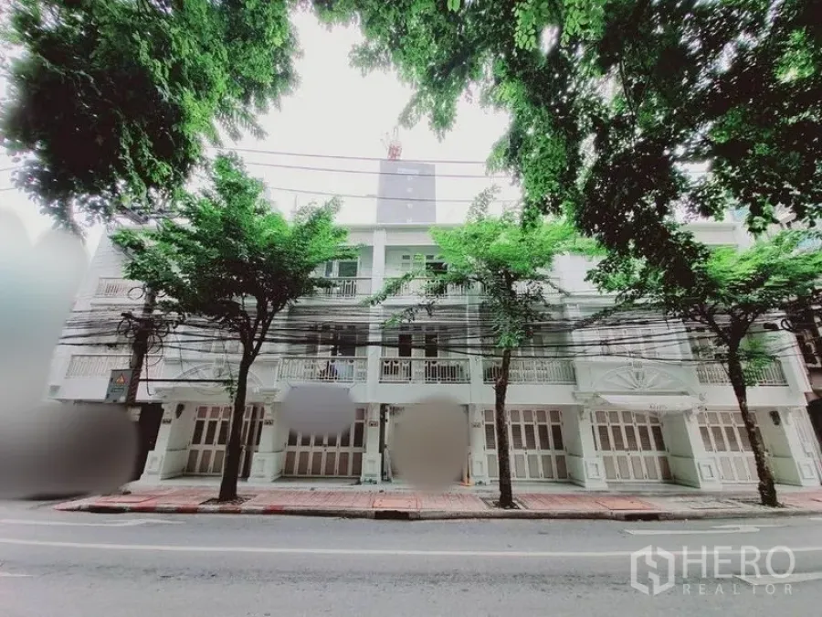 Shophouse for rent in Bang Rak, Bangkok - Tree-lined street view of a multi-storey shophouse facade with balconies and folding doors.