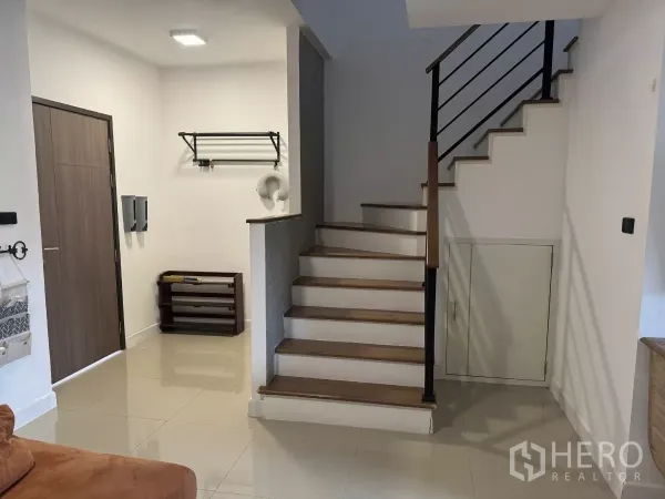 Townhouse for rent in Suan Luang, Bangkok - Entrance hall with wooden door, glossy tiles, and a staircase featuring a metal handrail and under‑stair storage.