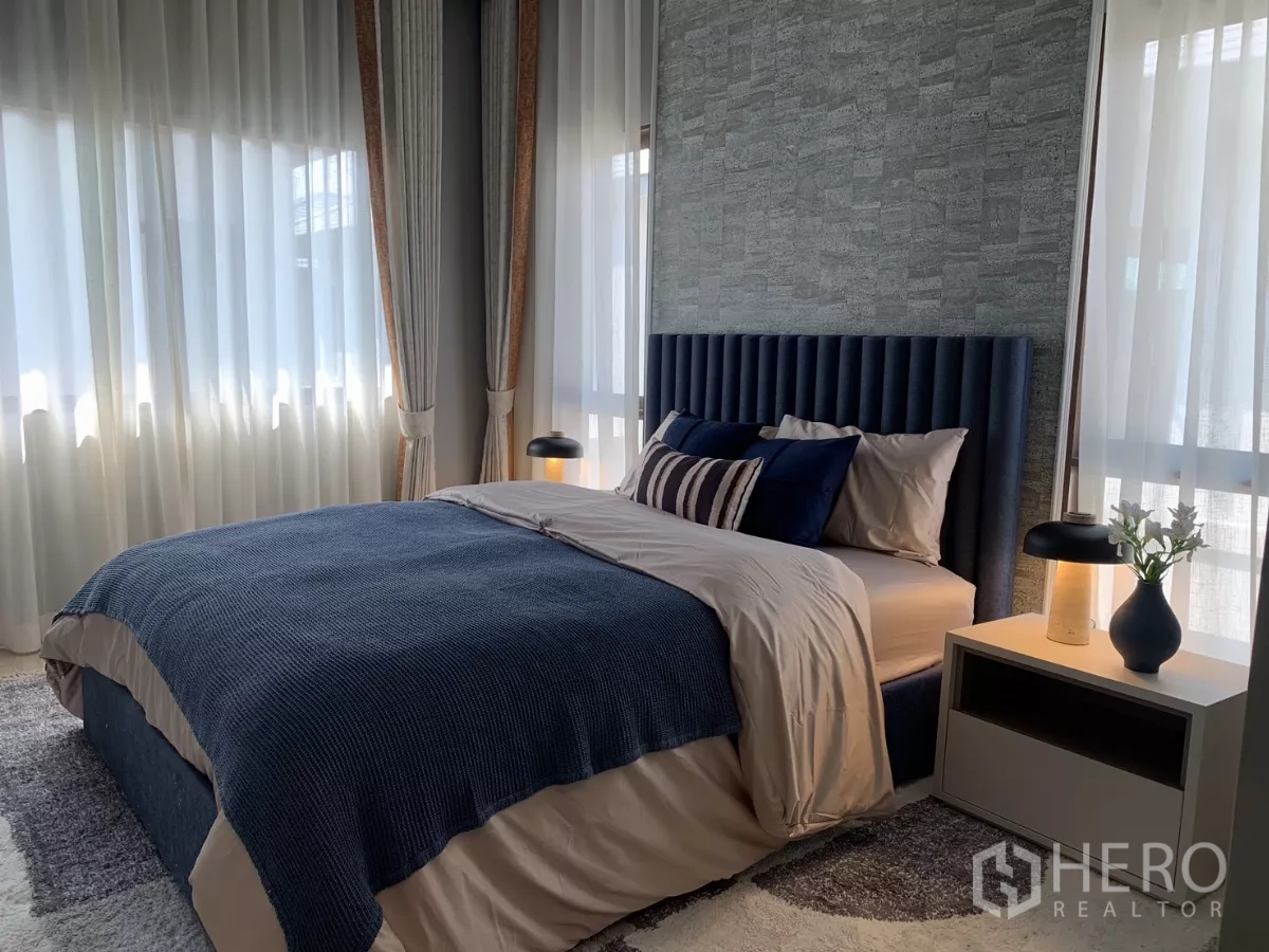 Detached House for rent in Bang Kapi, Bangkok - Sunlit bedroom with navy upholstered headboard, blue bedding and bedside lamp.