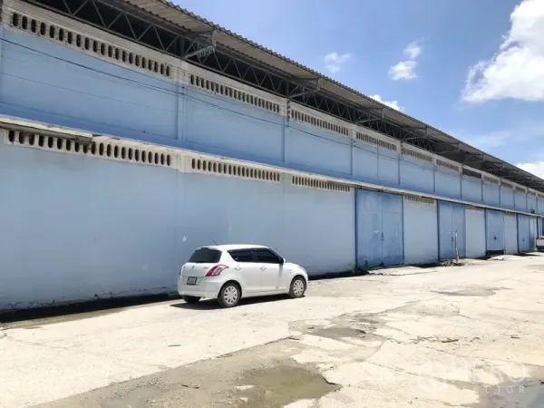 Warehouse for rent in Bang Phli, Samut Prakan - Side elevation of a long blue warehouse row with multiple loading gates and roadside parking under a clear sky.
