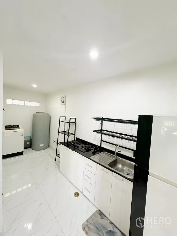 Townhouse for rent in Bang Phli, Samut Prakan - Fitted kitchen with black countertop, gas hob, sink, open shelving and an adjacent laundry zone.