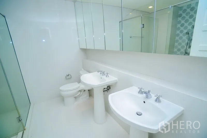 Condominium for rent in Khlong Toei, Bangkok - Large white bathroom featuring twin pedestal sinks, mirrored cabinets and a glass-enclosed shower.