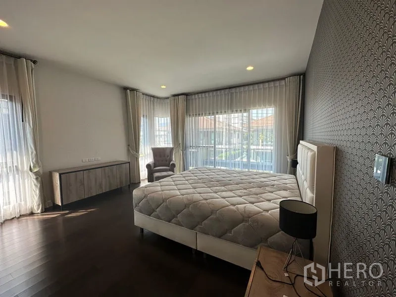 Detached House for rent or sale in Lam Luk Ka, Pathum Thani - Corner master bedroom with large windows, sheer curtains, feature wall, bed and armchair.