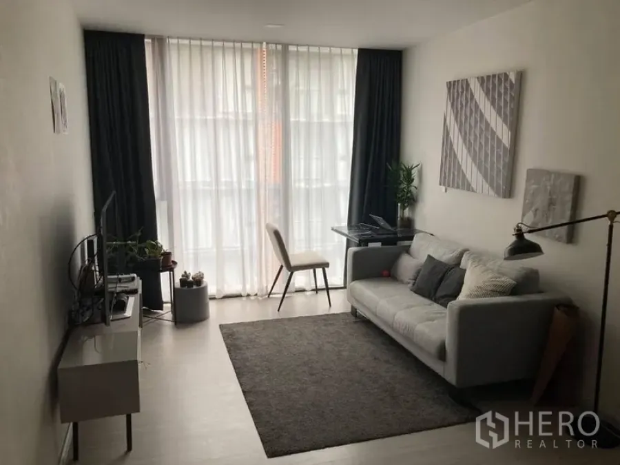 Condominium for rent in Khlong Toei, Bangkok - Bright living room with grey sofa, workspace and floor-to-ceiling curtains in a Bangkok condo.