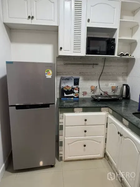 Townhouse for rent in Suan Luang, Bangkok - Compact L-shaped kitchen with white cabinets, stainless refrigerator, microwave, electric cooktop, and tiled backsplash.
