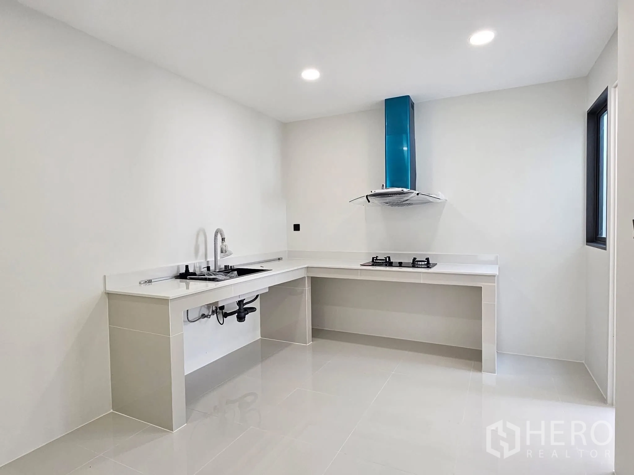 Detached House for sale in Lat Phrao, Bangkok - Minimalist white kitchen with L-shaped counters, dual gas hobs, and a stainless-steel hood.