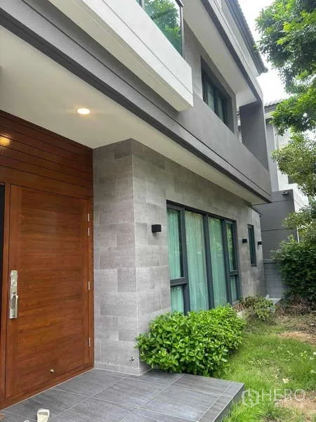 Detached House for rent in Mueang Nonthaburi, Nonthaburi - Front entrance showing solid wood door, grey stone cladding and low garden shrubs.