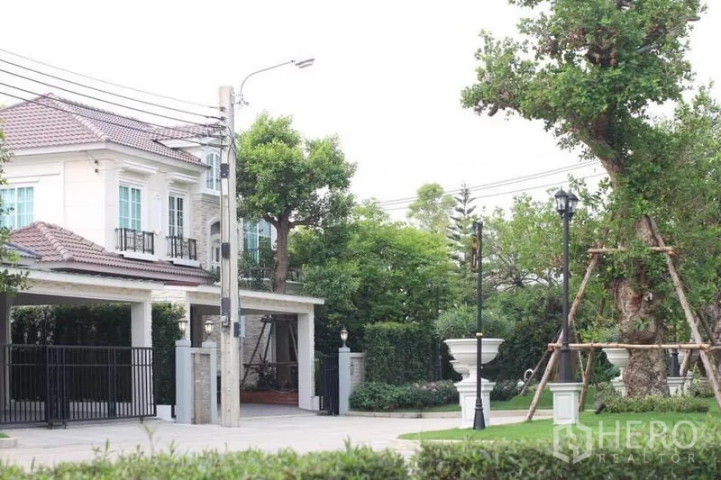 Detached House for rent or sale in Lat Phrao, Bangkok - Two-storey detached house with tiled roof, gated two-car carport and landscaped garden in a Bangkok neighborhood.