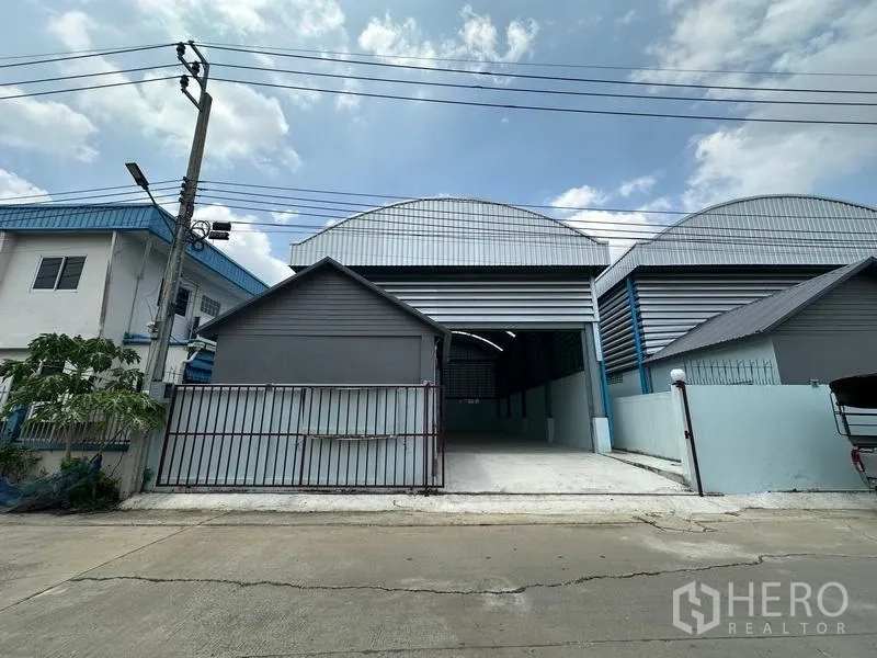 Factory for rent in Pak Kret, Nonthaburi - Front facade showing a tall roller shutter door and concrete driveway leading into the factory.