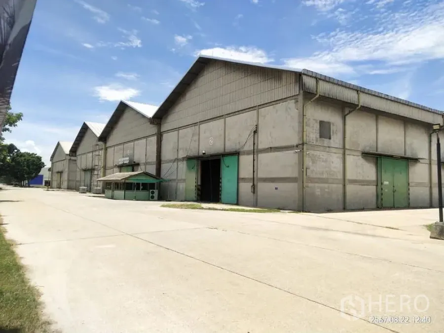 货栈 出租在Mueang Pathum Thani, Pathum Thani - 多栋人字形屋顶仓库，绿色推拉门，宽阔混凝土车道，蓝天下的巴吞他尼工业区景观。