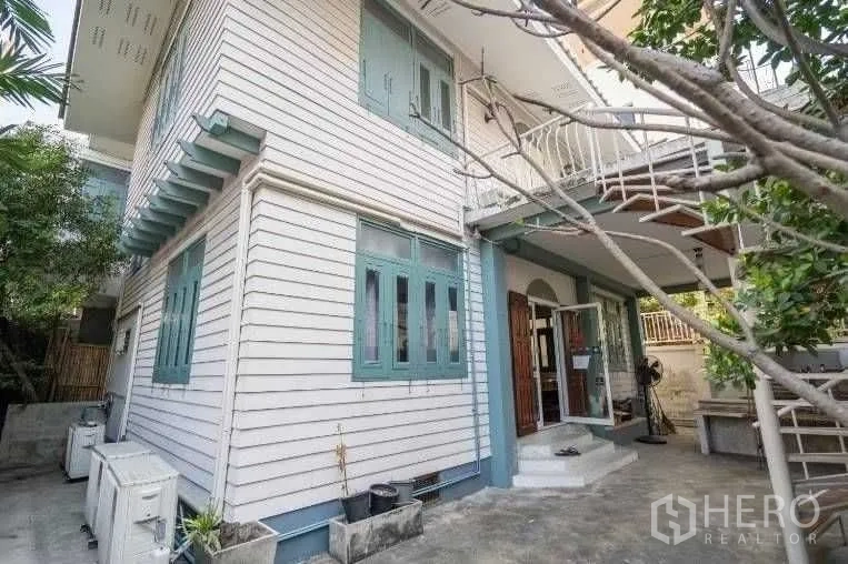 Detached House for rent in Khlong Toei, Bangkok - Two-storey detached house exterior with light cladding, green-framed windows, and a shaded patio with trees.