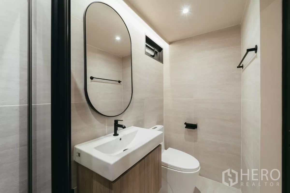 Townhouse for rent in Watthana, Bangkok - Powder room with oval mirror, wall-hung vanity and neutral tiles.