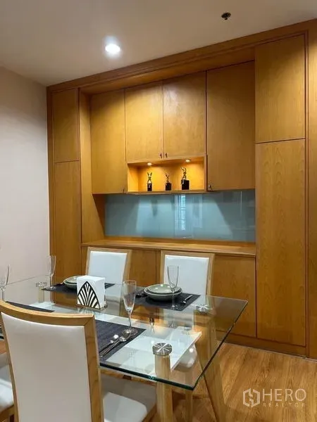 Condominium for rent in Khlong Toei, Bangkok - Dining area with glass-top table and four white chairs in front of built-in wood cabinetry with display shelf lighting.