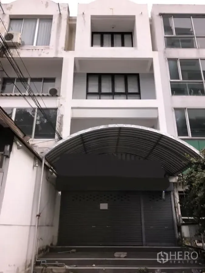 Townhouse for sale in Watthana, Bangkok - Four-storey white townhouse facade in Watthana, Bangkok, with roller shutter garage and curved metal awning.