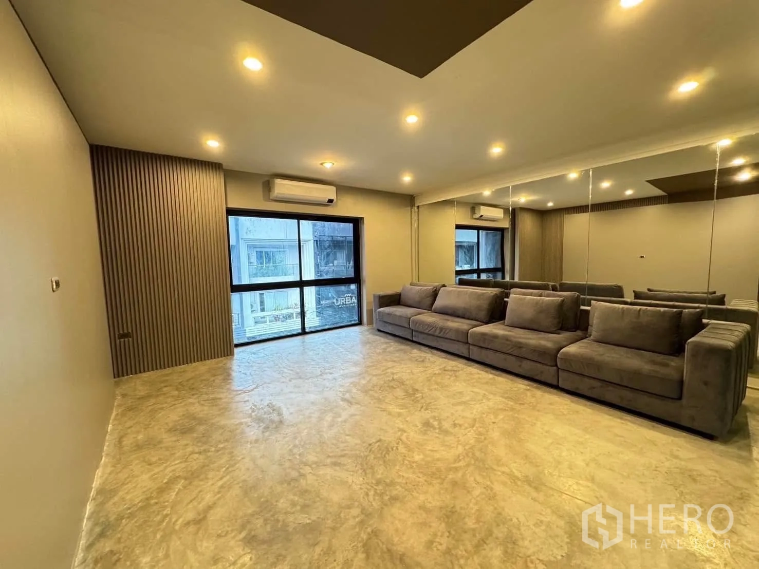 Home Office for rent in Watthana, Bangkok - Lounge with a long gray sofa, mirrored wall, recessed lighting and a large window.