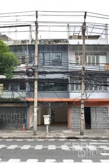 Shophouse for sale in Khlong Toei, Bangkok - Multi-storey shophouse facade on a main road in Khlong Toei, Bangkok, with metal roller shutters, open ground-floor bay, and overhead power lines.