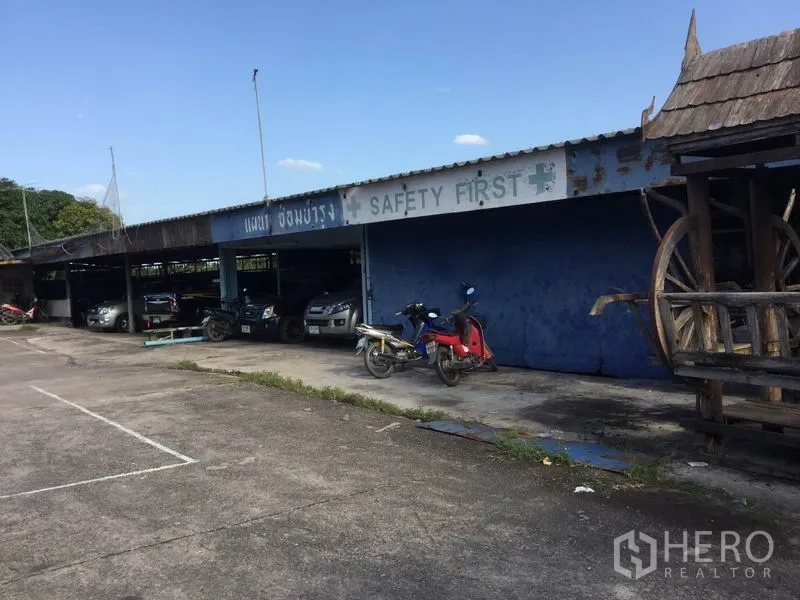 Warehouse for sale in Sai Noi, Nonthaburi - Sheltered parking bays with motorbikes and cars beside the yard, with signage on the canopy.