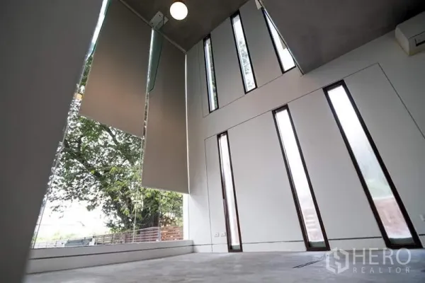 Townhouse for rent or sale in Phra Khanong, Bangkok - Double-height space with tall narrow windows and roller shades overlooking greenery.