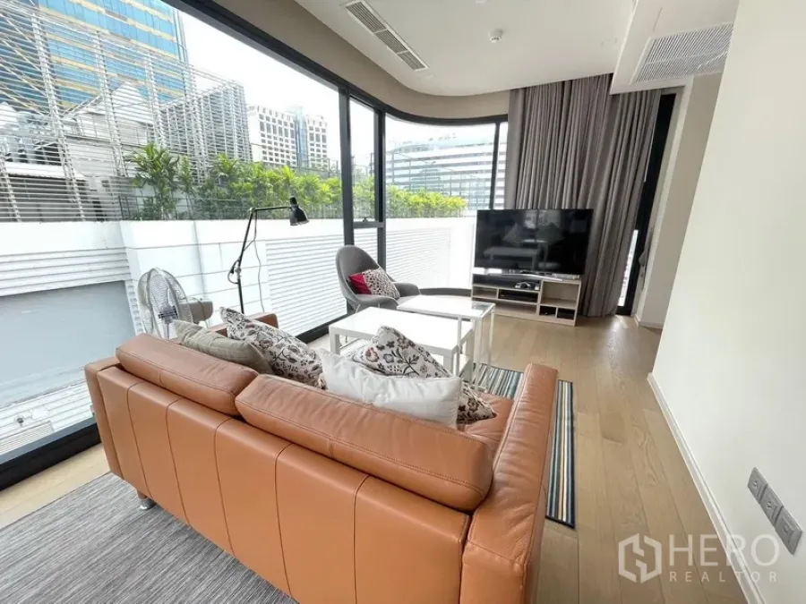 公寓 出租或出售在Watthana, Bangkok - 明亮客厅，棕色皮沙发、电视柜，转角落地窗可观绿意与城市景观。