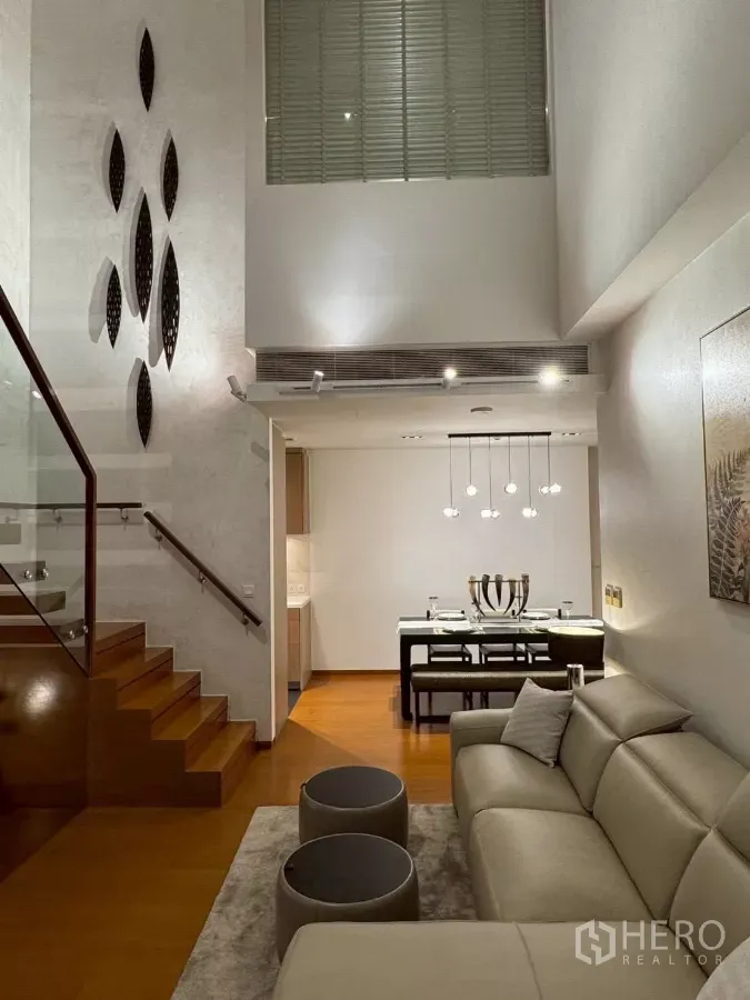 Condominium for rent in Sathon, Bangkok - Sathon luxury condo living and dining area with double-height ceiling, pendant lights, and wooden staircase with glass railing.